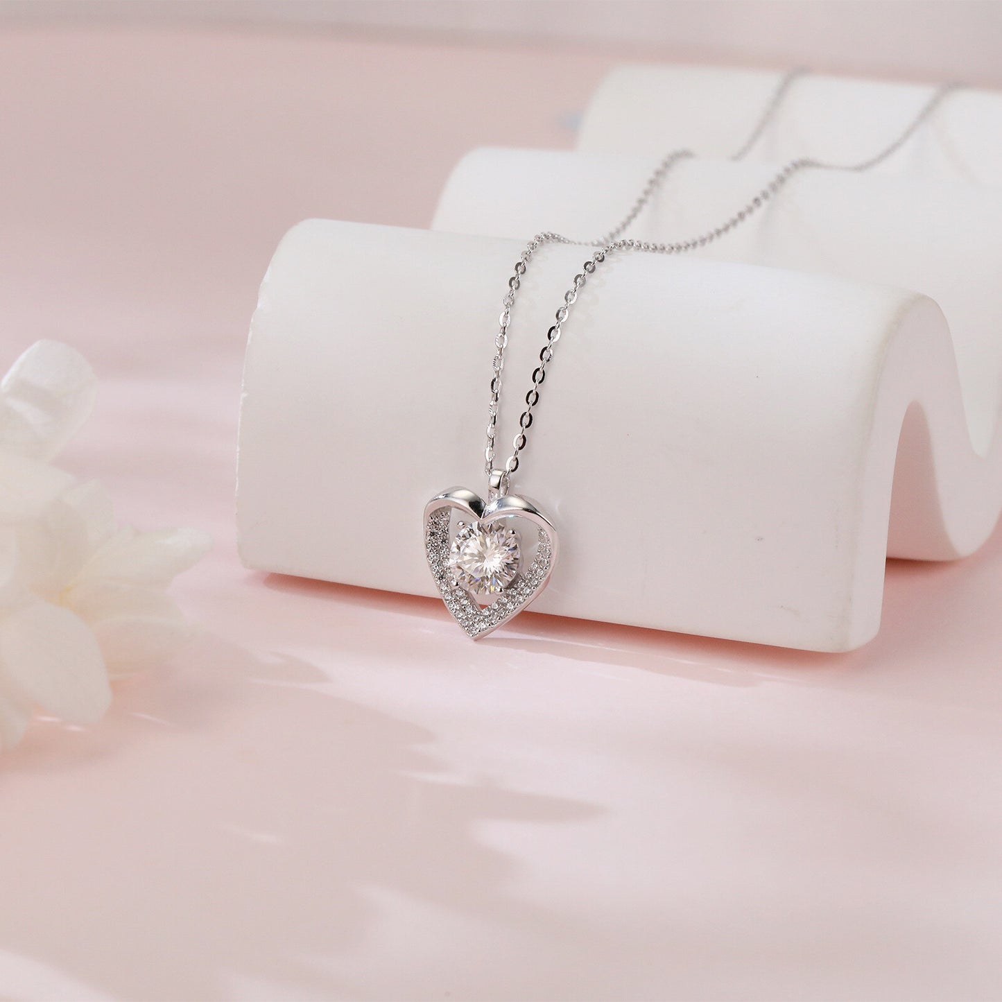 Heart-shaped silver necklace with clear stones on a white stand against a pink background