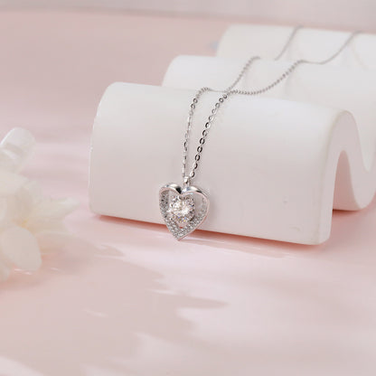 Heart-shaped silver necklace with clear stones on a white stand against a pink background