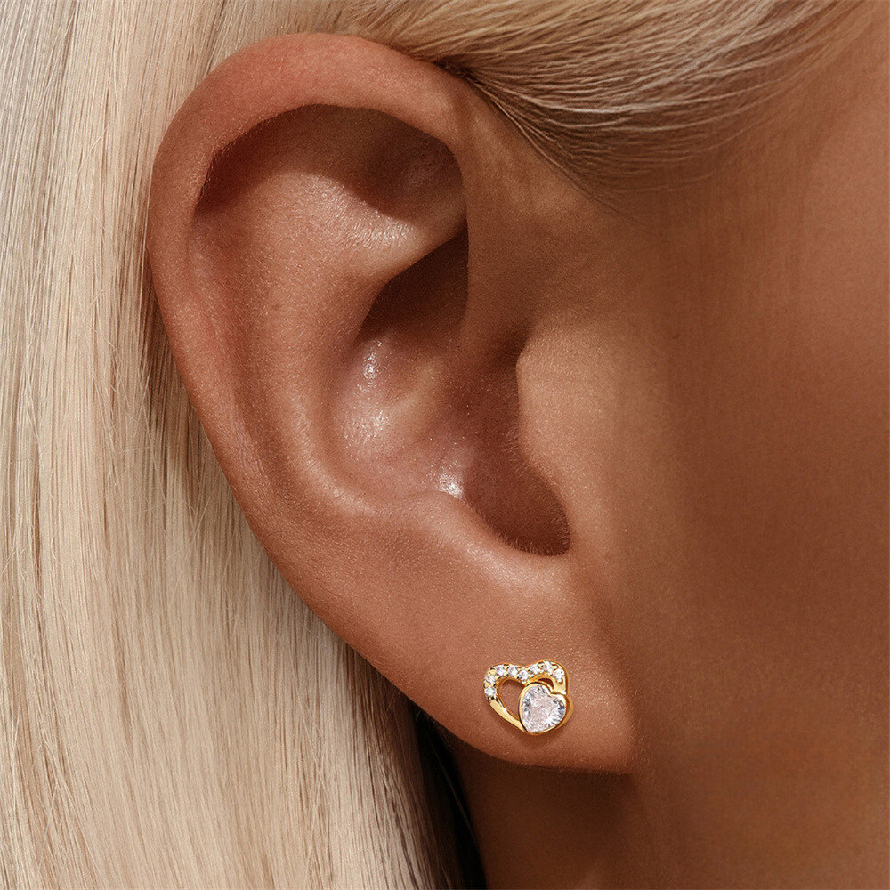 Close-up of an ear wearing a gold heart-shaped earring with a clear gemstone.