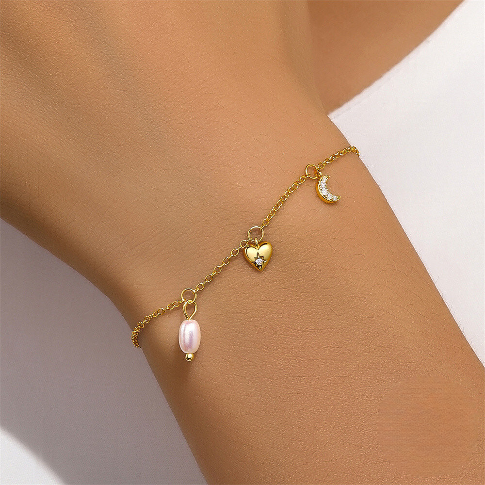 Gold bracelet with heart and pearl charm on a wrist against a neutral background