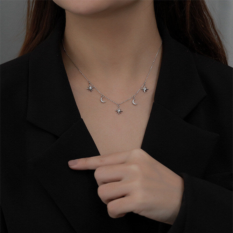 Person wearing a delicate necklace with star and moon charms, pointing to it.